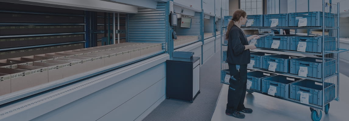 Woman picking orders into a mobile shelf of bins next to a row of vertical lift module (VLM) in a warehouse with gray overlay.