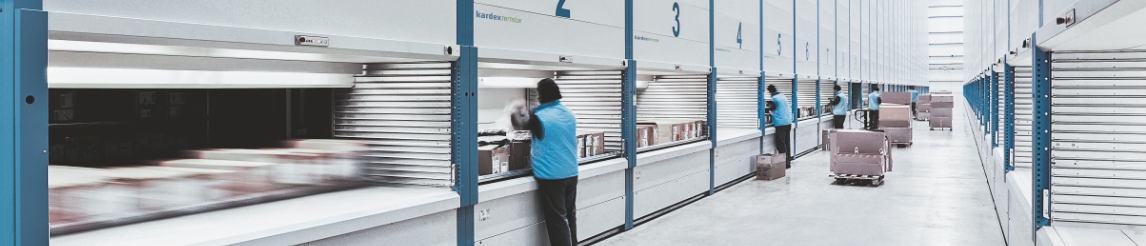 Worker in blue vest picking from a vertical lift module (VLM) in an aisle lined with additional VLMs. Additional workers and pallets in moving the background.