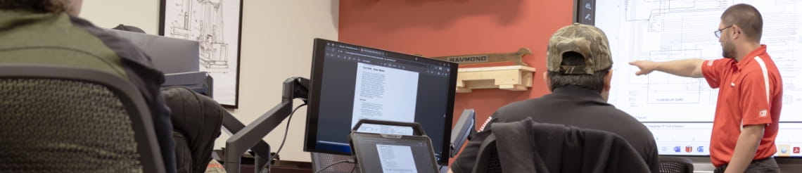RSC trainer in red at the front of a classroom pointing at a forklift schematic. Technician sitting at desk with computer looking at trainer.