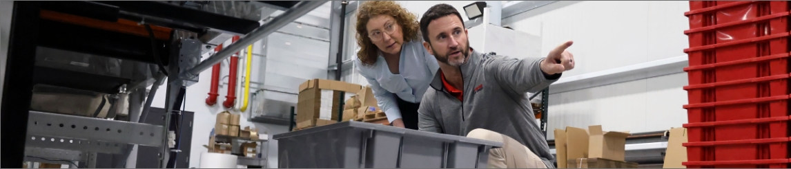 Male RSC engineer pointing down a conveyor with totes with a female customer behind him. She's looking where the engineer is pointing.