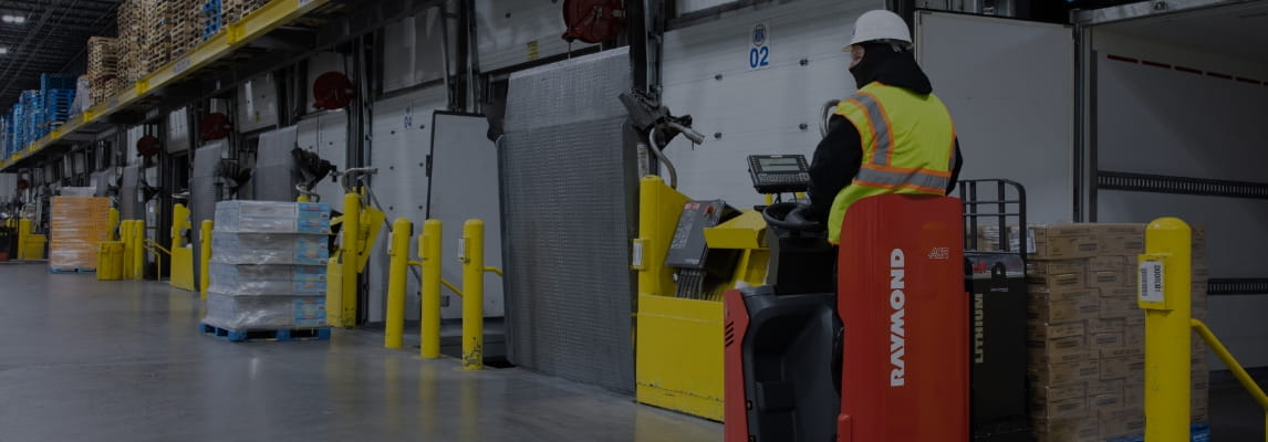 Forklift operator using a Raymond electric pallet jack to unload pallets from a trailer in a warehouse loading dock area.