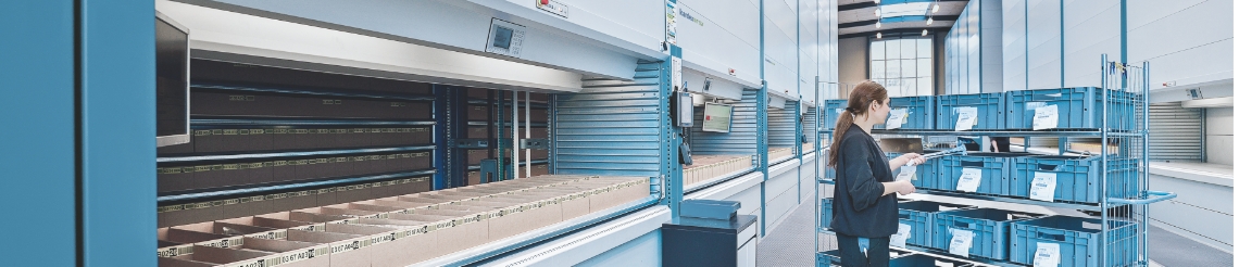 Woman picking orders into a mobile shelf of bins next to a row of vertical lift module (VLM) in a warehouse.