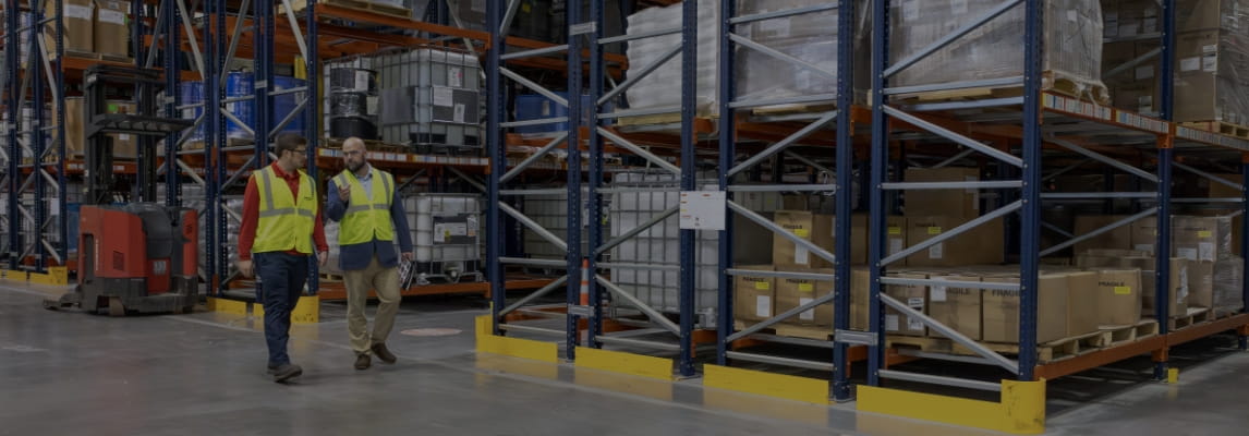 RSC rep and customer in safety vests talking and walking by aisles of warehouse racking with a Raymond forklift.