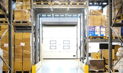A warehouse aisle with a dock door and air curtain at the end. Surrounded by racking full of boxes and pallets.