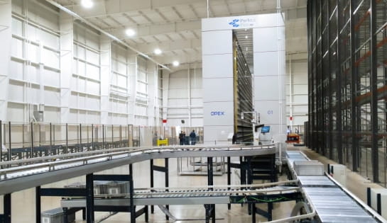 Totes moving around a conveyor that's entering and exiting a large OPEX Perfect Pick, an automated tote storage, inside a warehouse.