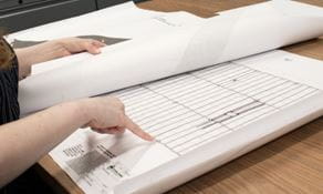 Design Engineer's hand pointing at warehouse blueprints on table.