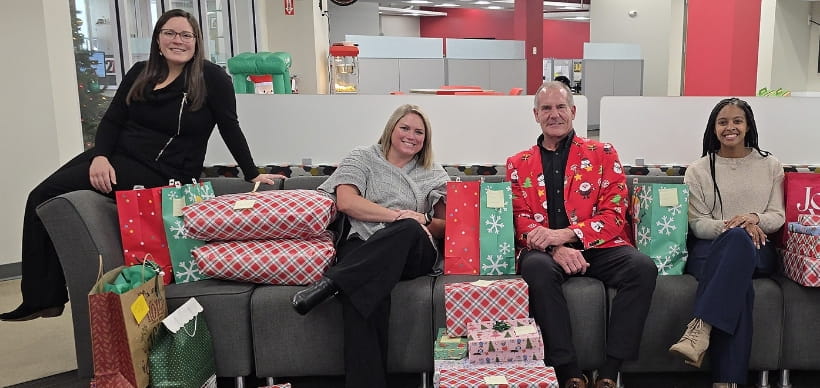 RSC Community Outreach team members posing with RSC President Scott Wolcott on couch with wrapped presents for Soldier's Angels Adopt-A-Family program.