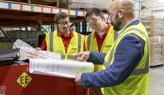 Two RSC employees standing next to a customer in a suit holding a blueprint open on top of a red Raymond forklift in a warehouse aisle.