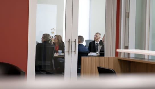 Group of people dressed in professional attire sitting at conference room table through glass doors.