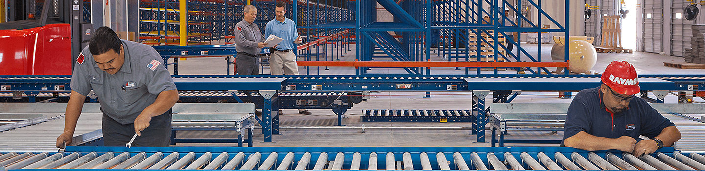 Raymond technicians maintaining a conveyor system with floor managers discussing plans in the background.