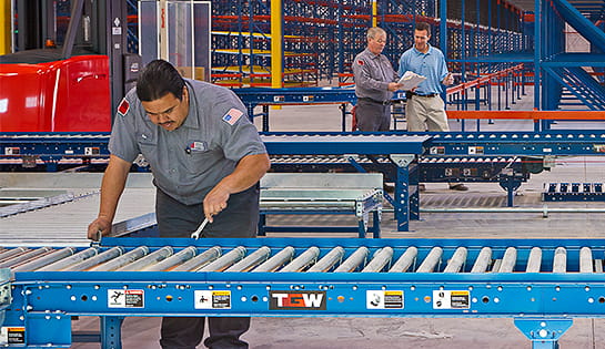 Technician using a wrench to install new conveyor equipment in warehouse. Two men in professional clothing talking in warehouse background.