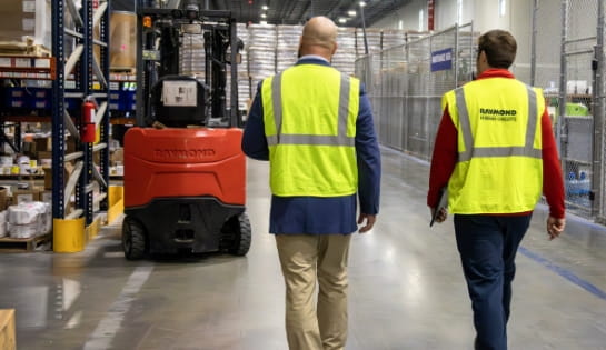 RSC employee and customer in suit jacket (both in safety vests) walking in a warehouse past multiple racking aisles and a Raymond forklift.