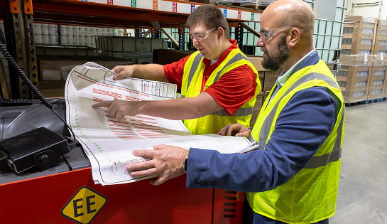 RSC engineer pointing a blueprint with an operation manager inside a warehouse racking aisle next to a Raymond forklift.