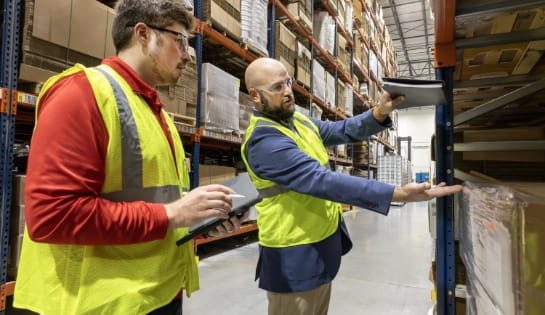 An operations manager is suit and safety vest measuring an empty space above a pallet with his hands. An RSC engineer listening beside the manager and taking notes.