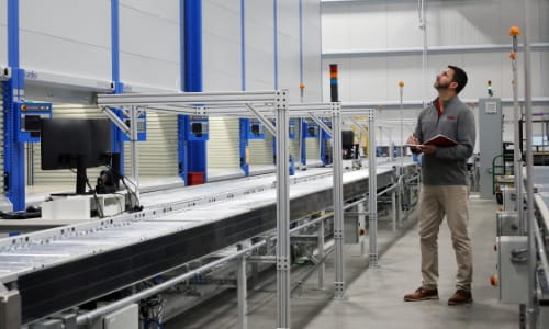 RSCI Sales Engineer looking up at warehouse space, observing the current system in place surrounded by an AS/RS system and conveyor. He's holding a notebook with pen for notes.