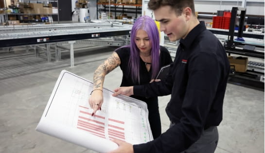 Female RSC project manager pointing at a racking blueprint being held by male RSC engineer in a warehouse. Conveyor in the background.
