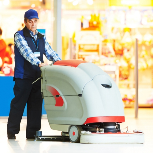 Man in cap and vest pushing a gray and red floor scrubber in retail space.