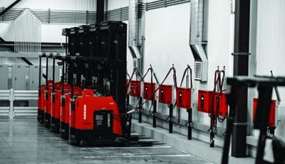 Warehouse wall with several red battery chargers hung. A row of Raymond reach trucks lined up charging.