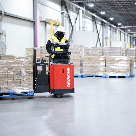Warehouse worker driving a red Raymond electric pallet jack with loaded pallet to warehouse staging area.