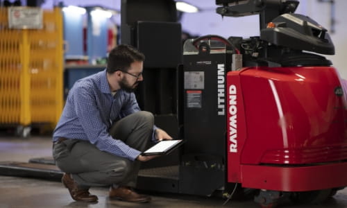 Man in professional attire squat down with tablet inspecting Raymond forklift.
