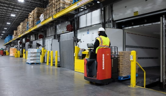 Forklift operator using a Raymond electric pallet jack to unload pallets from a trailer in a warehouse loading dock area.