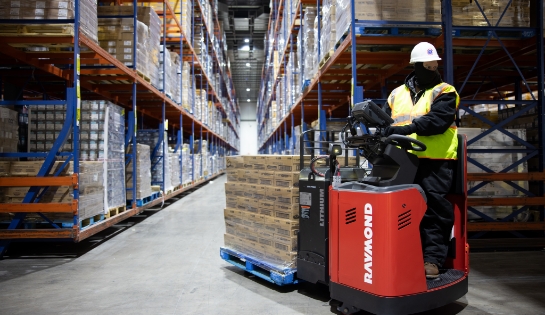Warehouse worker driving a Raymond forklift towing a pallet of boxes through a racking aisle. Worker is wearing a safety vest, hard hat and face mask.