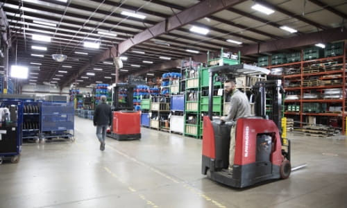 Worker in Raymond counterbalanced forklift crossing pedestrian walkway in warehouse. Pedestrian walking in marked footpath. Racking with pallets in background.