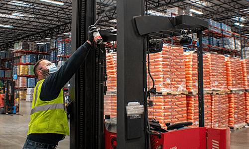 Raymond Storage Concepts technician in safety vest spraying lubricant on Raymond forklift during maintenance service.