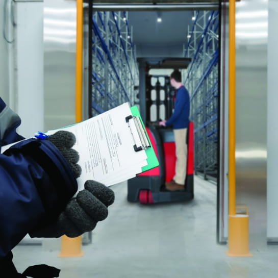 Raymond forklift going into a warehouse freezer room with a gloved hand holding a clipboard with papers in the foreground.