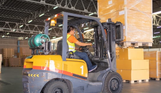 Yellow LP gas powered forklift lifting a wrapped pallet of boxes. Driver is moving the pallet through a warehouse staging area.