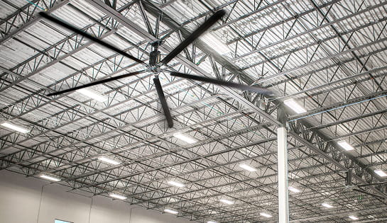 Looking up at an industrial ceiling fan in a warehouse, surrounded by rafters.