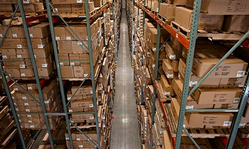 Narrow warehouse aisle lined on either side with green and red racking filled with pallets and boxes.