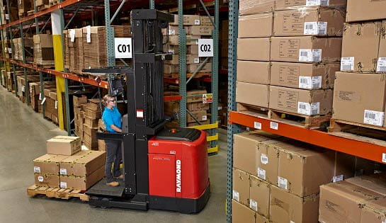 Woman driving a Raymond orderpicker with pallet of boxes on the back. Driving through warehouse aisles of racking.