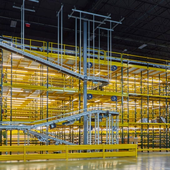 Yellow picking module in warehouse with three levels. Conveyor going along the side stopping at each level to pick up orders.