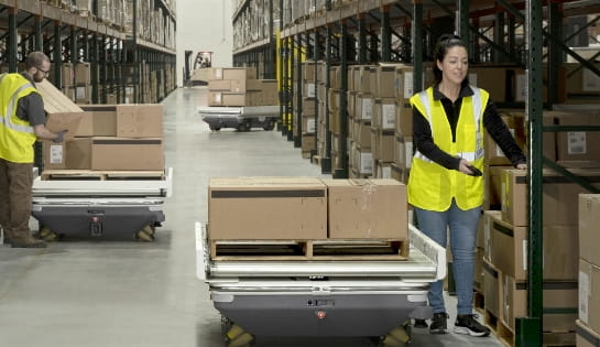 Warehouse worker picking orders and placing them on large robot with pallet in warehouse aisle.