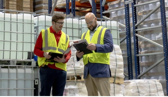 Man in suite jacket and safety vest looking RSC marketing materials while talking to RSC rep in red and safety vest. Both are standing at the end of warehouse racking aisle with pallets behind them.