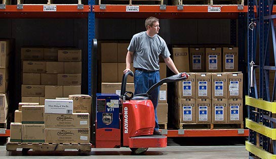 Warehouse worker riding a Raymond end rider pallet jack with a pallet of boxes through a warehouse aisle.