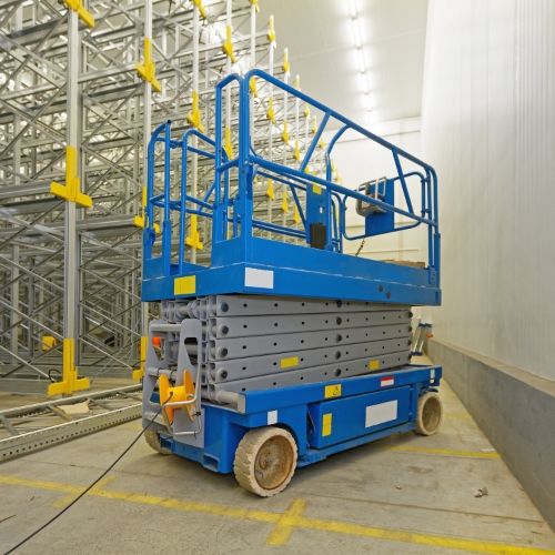 Blue scissor lift parked in warehouse with metal racking behind it.