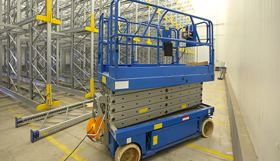 Blue scissor lift parked in warehouse with metal racking behind it.