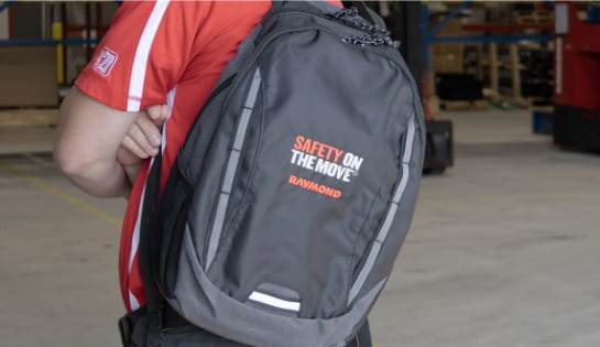 Person in red shirt holding a Raymond Safety On The Move black backpack on his back in a warehouse.