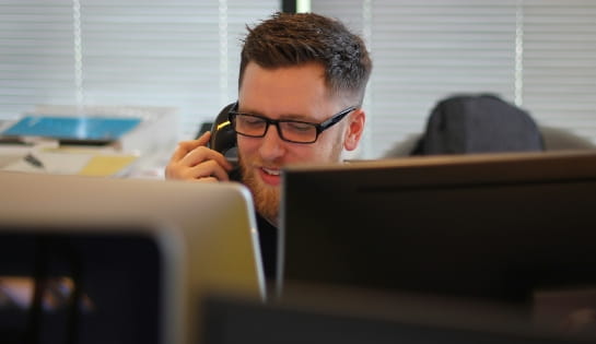 man with glasses talking on the phone in the office, seen over the top of computer monitors.