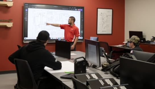 Trainer in front of a classroom of technicians pointing at a forklift schematic on a TV screen. Technicians sitting at desks with computers and monitors.