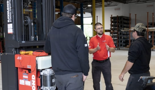 Trainer in red RSC polo talking to two technicians during hands-on portion of training in warehouse next to opened forklift engine.