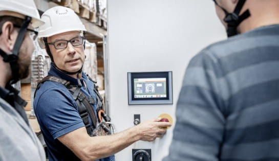 Man in a hard hat showing two other people how the controllers and monitor work on a piece of equipment.