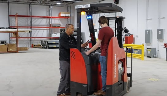 Trainer in black RSC jacket talking with trainee in red shirt about forklift controls while he's standing in a counterbalanced lift truck. They are in the middle of a warehouse training floor surrounded by racking and boxes.