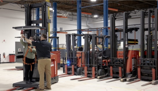 Trainer in black polo demonstrating a fall harness pulley to forklift operator trainee that is standing on a orderpicker in warehouse with a line of other forklifts.