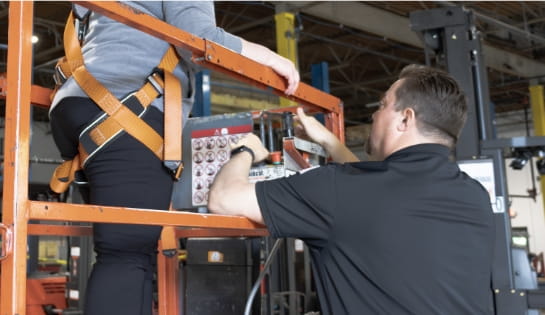 Trainer showing operator how a scissor lift's controls work. Operator is in a safety harness standing on scissor lift.