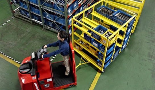 Female warehouse worker driving a Raymond tugger with attached carts.