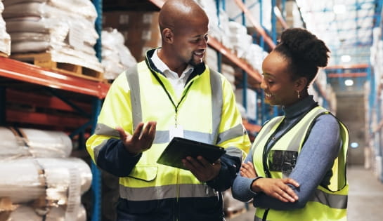 Two warehouse workers wearing safety vests looking at a tablet in a racking aisle.