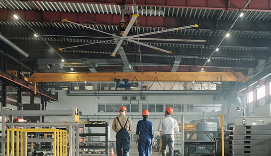 Three hard-hatted workers walking in a warehouse with an industrial ceiling fan above.